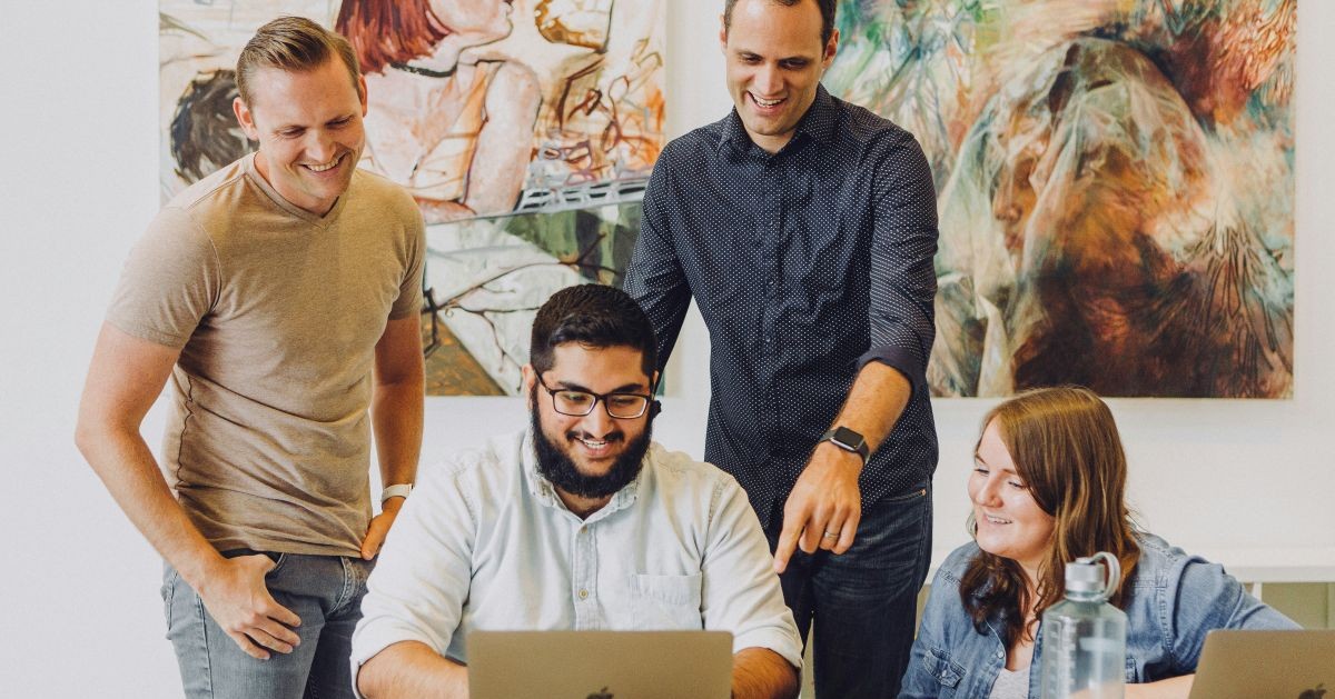 People stood around one person on a laptop. They are smiling and working together.
